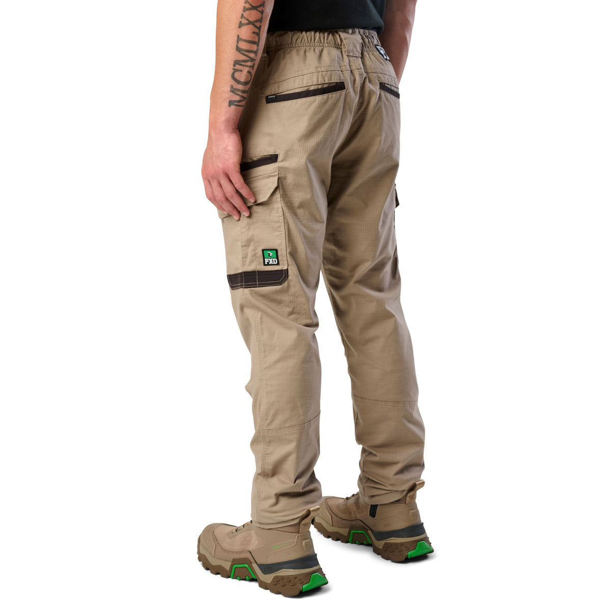 A 3-4 back view of a man wearing khaki cargo style FXD Men's WP-12 Elastic Waist Stretch Ripstop Work Pants (FX02506026) with stone coloured work boots on a white background.