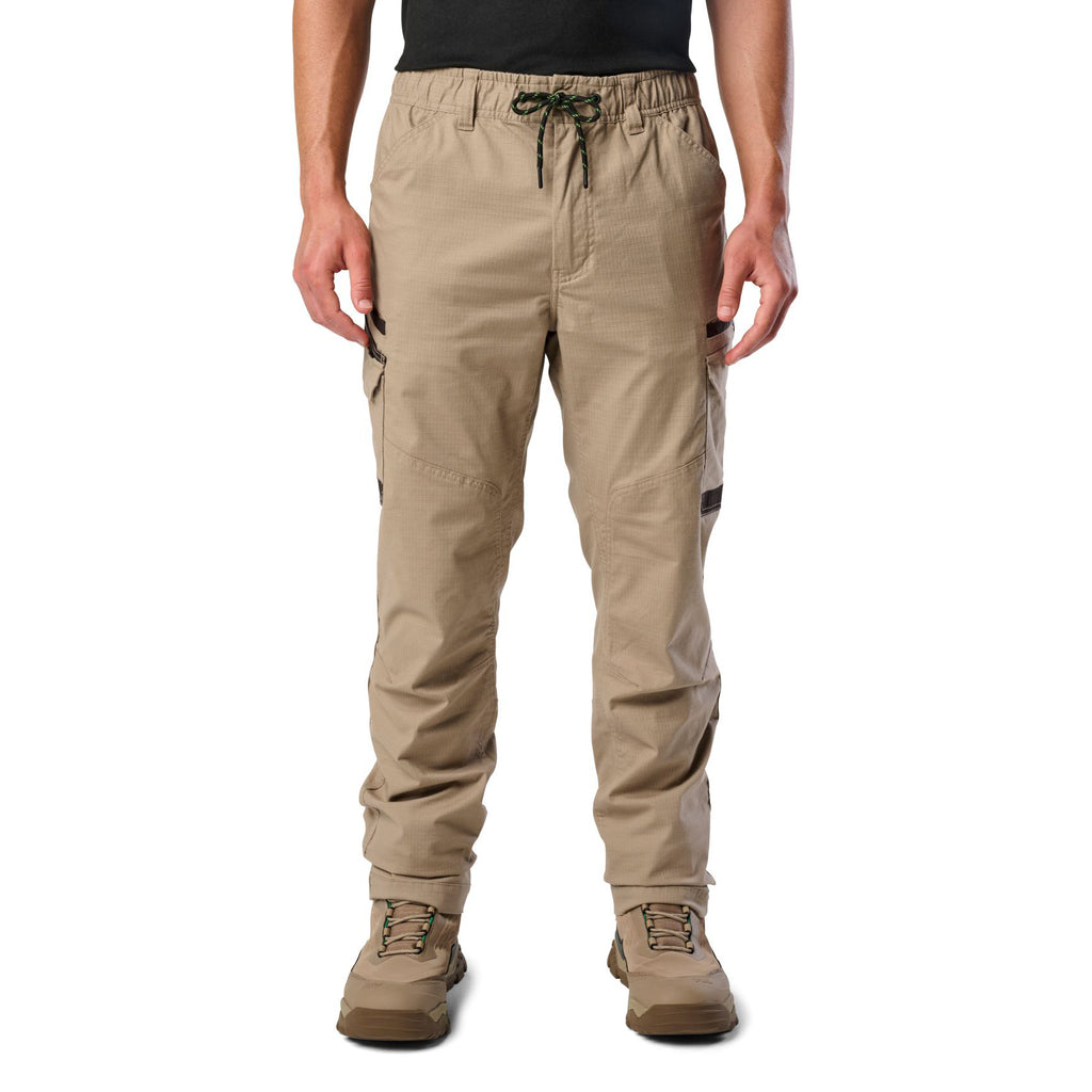 A front view of a man wearing khaki cargo style FXD Men's WP-12 Elastic Waist Stretch Ripstop Work Pants (FX02506026) with stone coloured work boots on a white background.