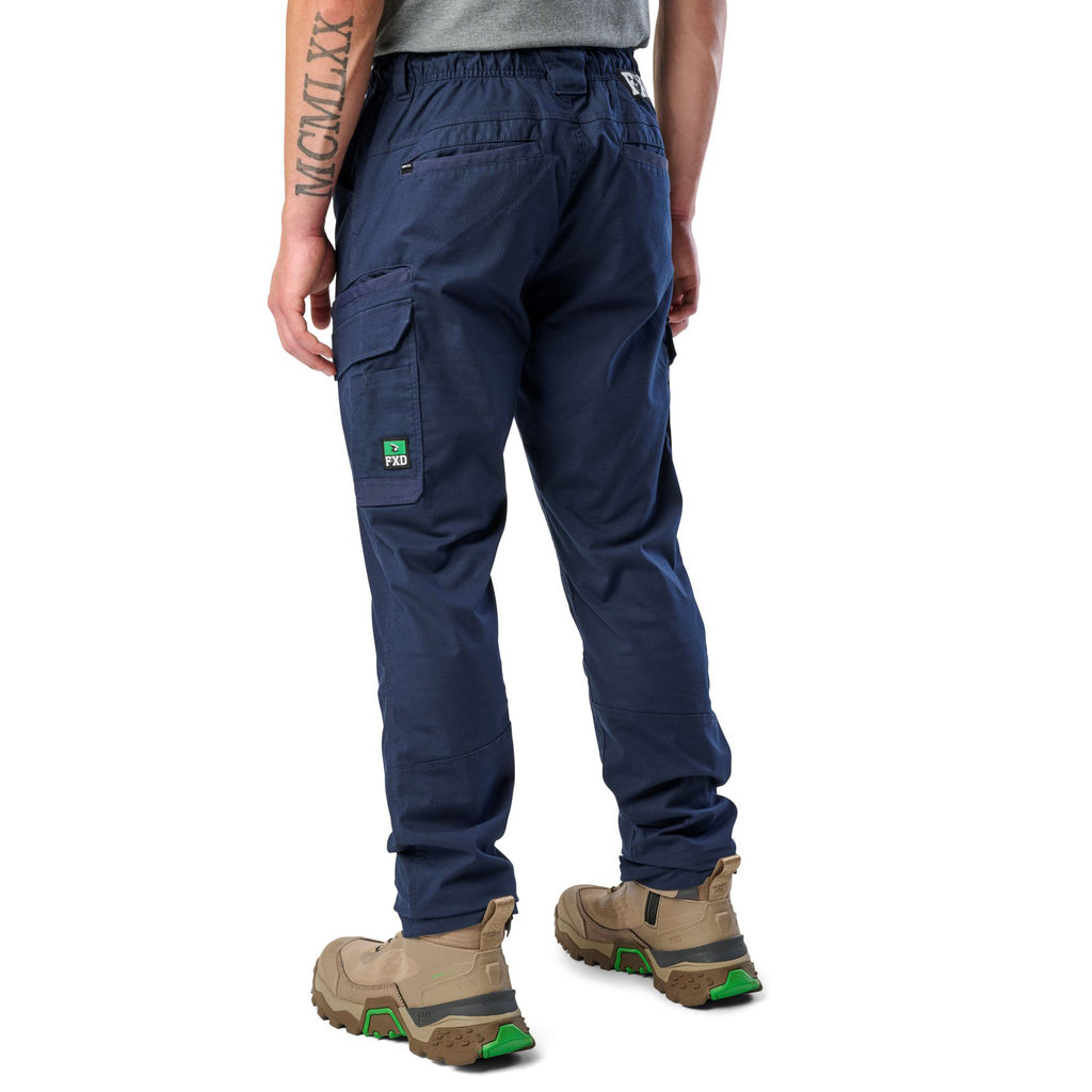 A 3-4 back view of a man wearing navy blue cargo style FXD Men's WP-12 Elastic Waist Stretch Ripstop Work Pants (FX02506026) with stone coloured work boots on a white background.