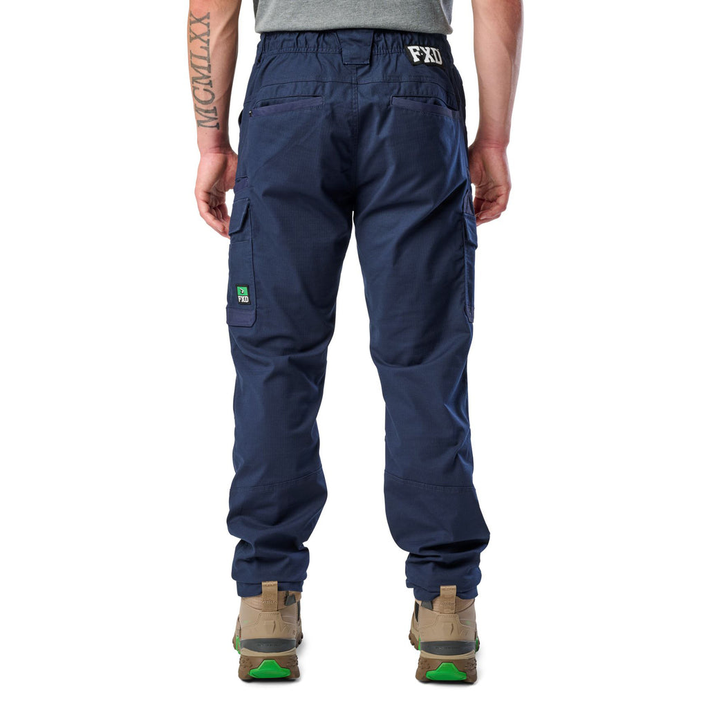A back view of a man wearing navy blue cargo style FXD Men's WP-12 Elastic Waist Stretch Ripstop Work Pants (FX02506026) with stone coloured work boots on a white background.
