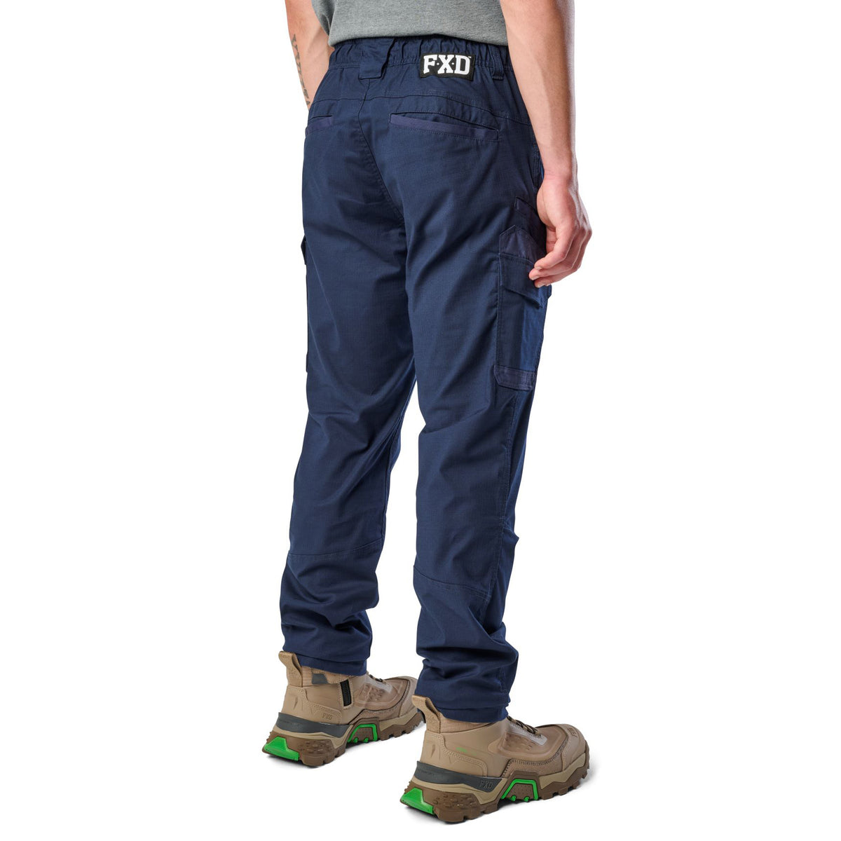 A 3-4 back view of a man wearing navy blue cargo style FXD Men's WP-12 Elastic Waist Stretch Ripstop Work Pants (FX02506026) with stone coloured work boots on a white background.