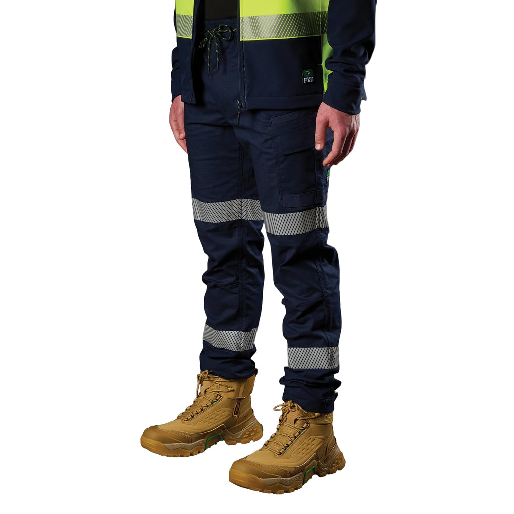 A 3-4 front view of a man wearing a pair of navy blue FXD Men's WP-12T Elastic Waist Stretch Ripstop Taped Work Pants (FX02606028) with a hi vis jacket and wheat coloured work boots, on a white background.