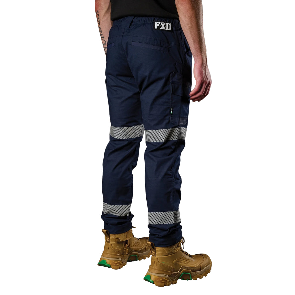A 3-4 back view of a man wearing a pair of navy blue FXD Men's WP-12T Elastic Waist Stretch Ripstop Taped Work Pants (FX02606028) with a black tee and wheat coloured work boots, on a white background.