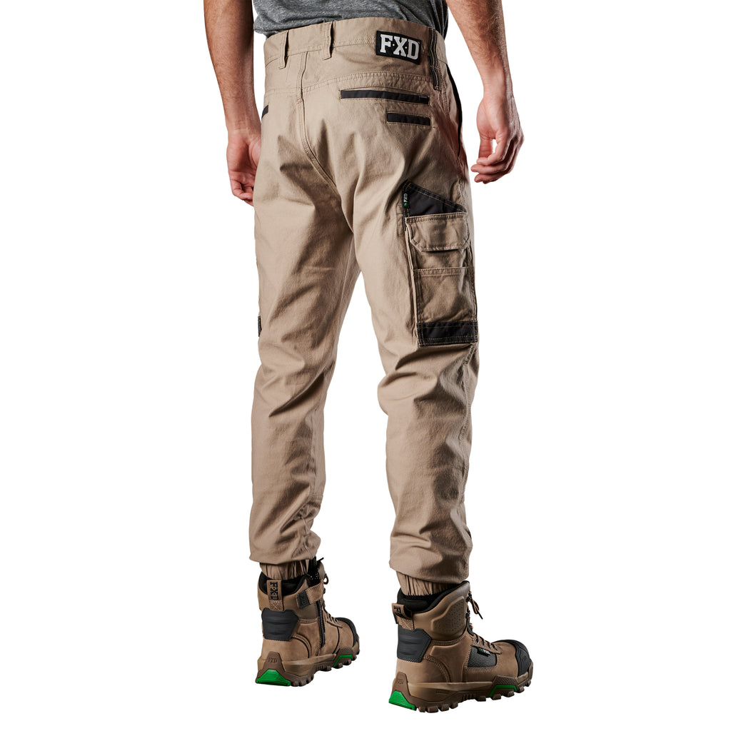 A 3-4 back view of a man wearing khaki FXD Men's WP-4 Stretch Cuffed Work Pants (FX01616003) with cargo pockets and elastic cuffed ankles, paired with wheat coloured work boots, shown against a plain white background.