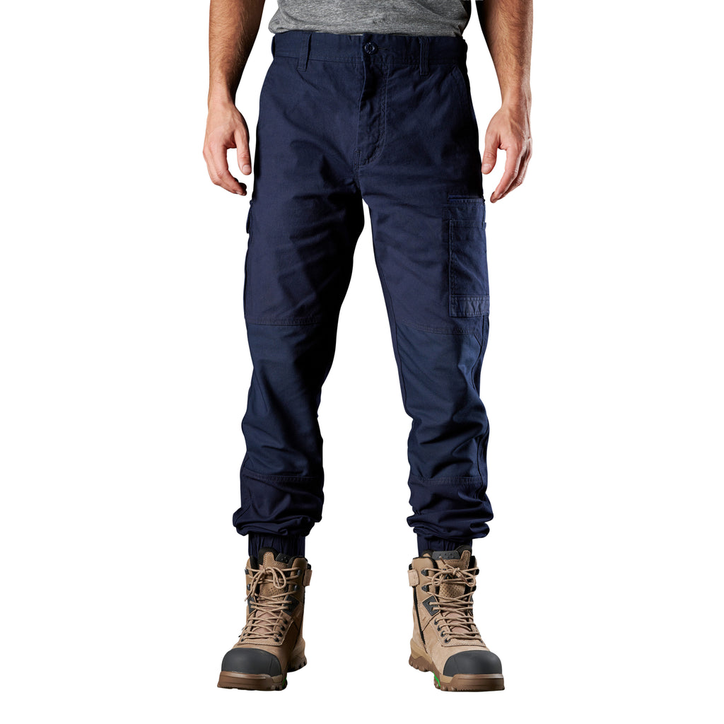 A man wearing navy FXD Men's WP-4 Stretch Cuffed Work Pants (FX01616003) with cargo pockets and elastic cuffed ankles, paired with wheat coloured work boots, shown against a plain white background.