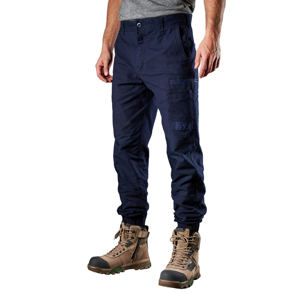 A 3-4 front view of a man wearing navy FXD Men's WP-4 Stretch Cuffed Work Pants (FX01616003) with cargo pockets and elastic cuffed ankles, paired with wheat coloured work boots, shown against a plain white background.