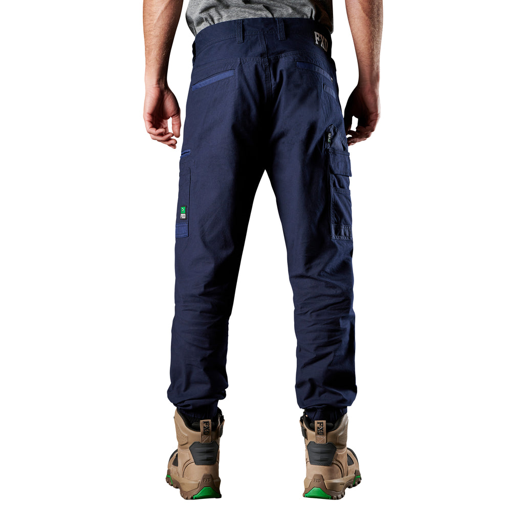 A back view of a man wearing navy FXD Men's WP-4 Stretch Cuffed Work Pants (FX01616003) with cargo pockets and elastic cuffed ankles, paired with wheat coloured work boots, shown against a plain white background.