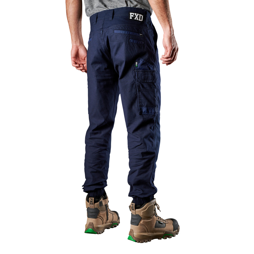 A 3-4 back view of a man wearing navy FXD Men's WP-4 Stretch Cuffed Work Pants (FX01616003) with cargo pockets and elastic cuffed ankles, paired with wheat coloured work boots, shown against a plain white background.