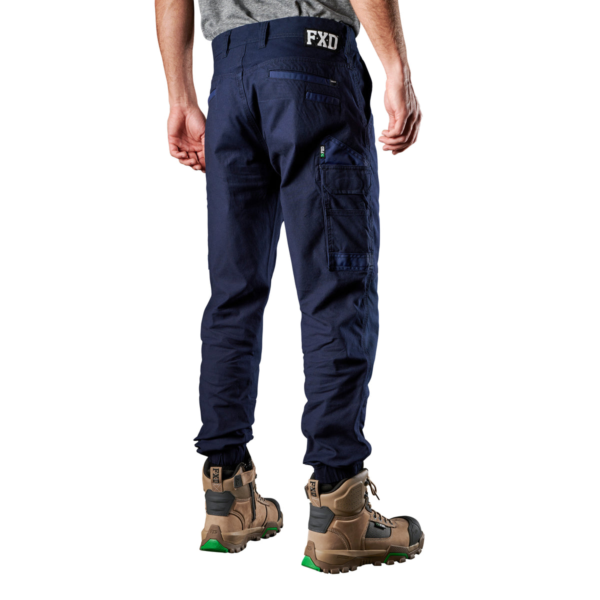 A 3-4 back view of a man wearing navy FXD Men's WP-4 Stretch Cuffed Work Pants (FX01616003) with cargo pockets and elastic cuffed ankles, paired with wheat coloured work boots, shown against a plain white background.