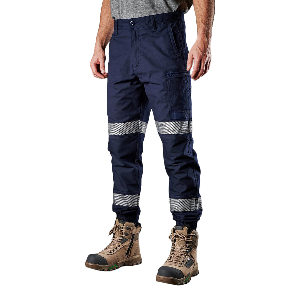 A 3-4 front view of a man wearing navy blue FXD Men's WP-4T Reflective Cuffed Work Pants (FX02106017) with reflective silver bands on the legs, paired with a grey t-shirt and stone coloured work boots, against a plain white background.