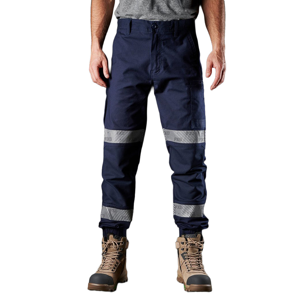 A man wearing navy blue FXD Men's WP-4T Reflective Cuffed Work Pants (FX02106017) with reflective silver bands on the legs, paired with a grey t-shirt and stone coloured work boots, against a plain white background.