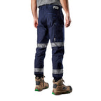 A 3-4 back view of a man wearing navy blue FXD Men's WP-4T Reflective Cuffed Work Pants (FX02106017) with reflective silver bands on the legs, paired with a grey t-shirt and stone coloured work boots, against a plain white background.