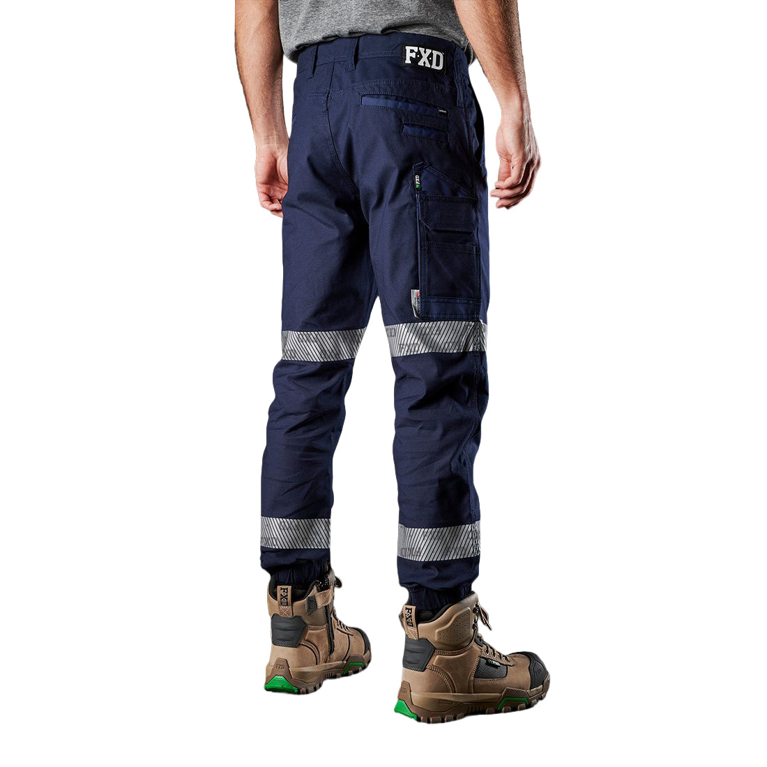A 3-4 back view of a man wearing navy blue FXD Men's WP-4T Reflective Cuffed Work Pants (FX02106017) with reflective silver bands on the legs, paired with a grey t-shirt and stone coloured work boots, against a plain white background.