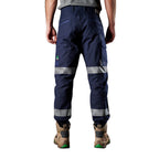 A back view of a man wearing navy blue FXD Men's WP-4T Reflective Cuffed Work Pants (FX02106017) with reflective silver bands on the legs, paired with a grey t-shirt and stone coloured work boots, against a plain white background.