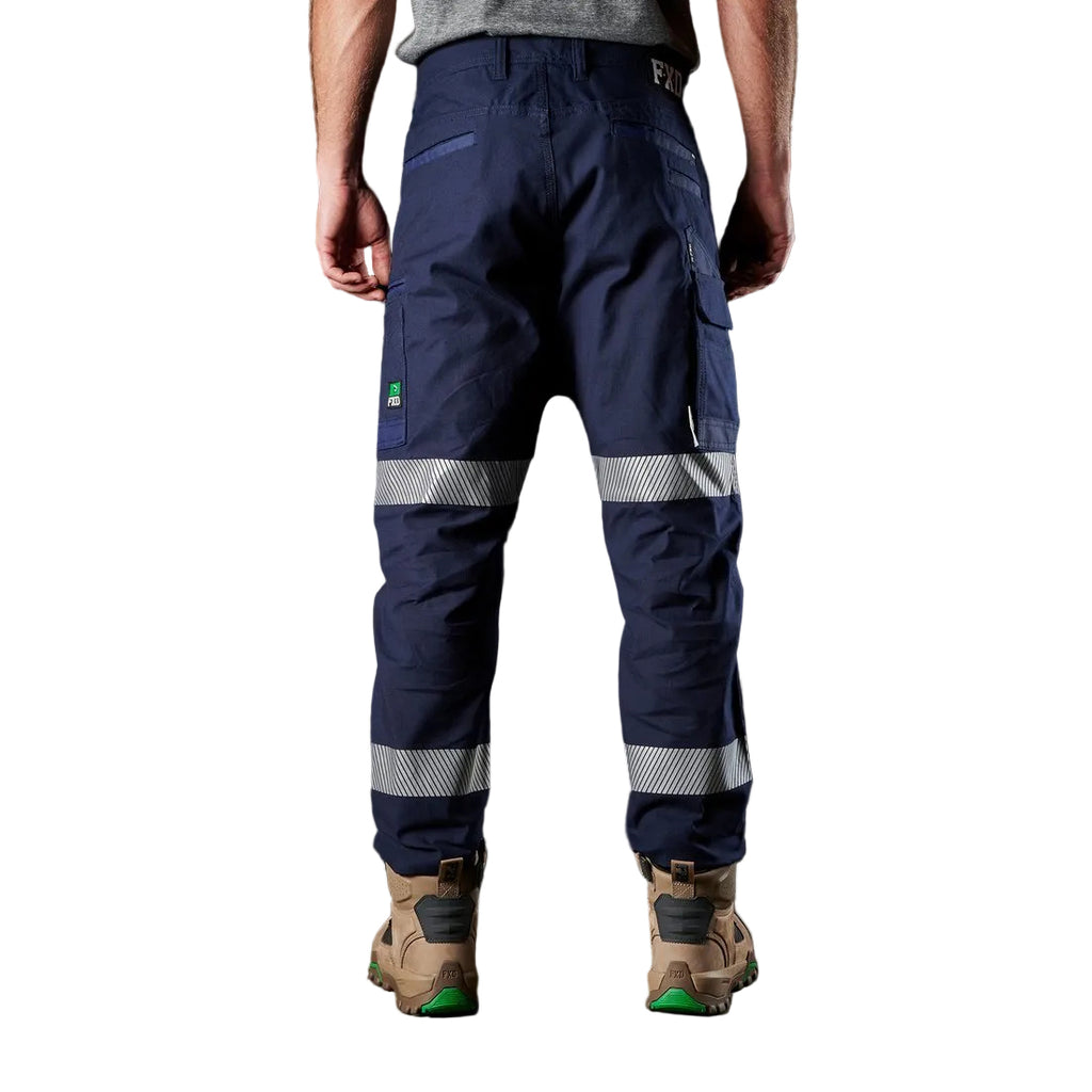 A model from the back, from the waist down, wearing navy blue FXD WP-3T Reflective Stretch Work Pants (FX01906010) with reflective silver stripes on the thighs and lower legs, paired with stone coloured work boots against a white background.