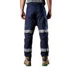 A model from the back, from the waist down, wearing navy blue FXD WP-3T Reflective Stretch Work Pants (FX01906010) with reflective silver stripes on the thighs and lower legs, paired with stone coloured work boots against a white background.