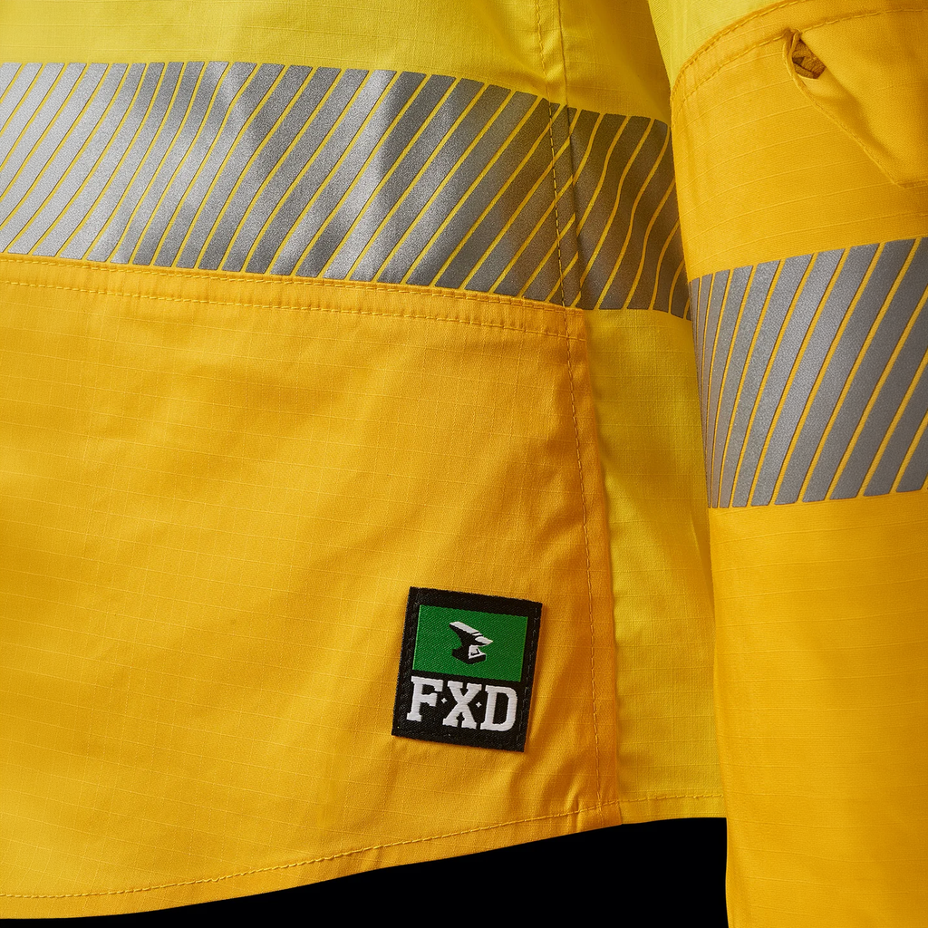 Close up detail of tape and the FXD logo badge at the hem on a yellow FXD Women's LSH-2WT Hi Vis Taped Long Sleeve Work Shirt (FX12404100) on a white background.