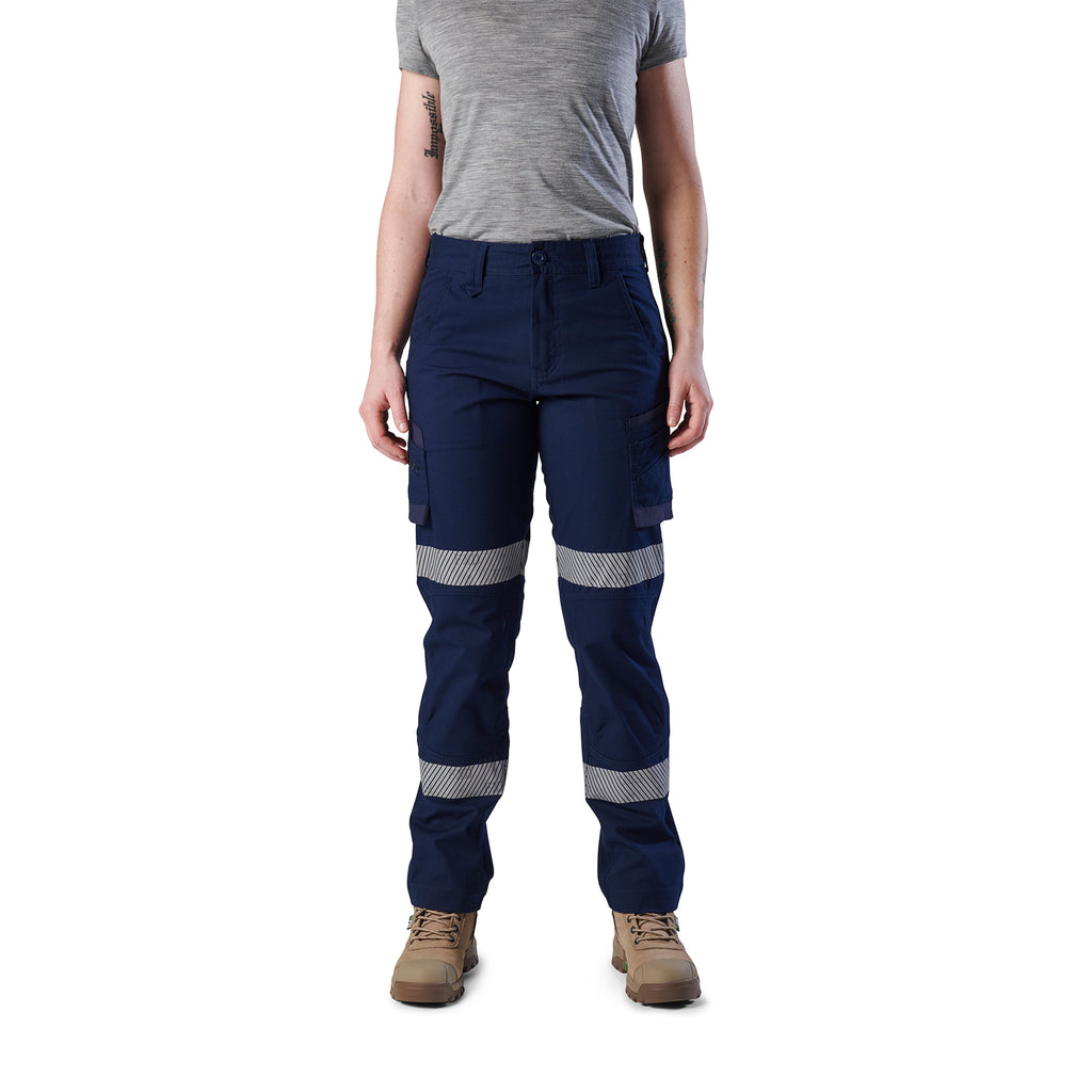 A woman wearing navy blue FXD Women's Taped Cargo Work Pants (WP-7WT) with reflective silver bands on the legs, paired with a grey t-shirt and stone coloured work boots, against a plain white background.