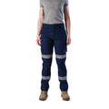 A woman wearing navy blue FXD Women's Taped Cargo Work Pants (WP-7WT) with reflective silver bands on the legs, paired with a grey t-shirt and stone coloured work boots, against a plain white background.