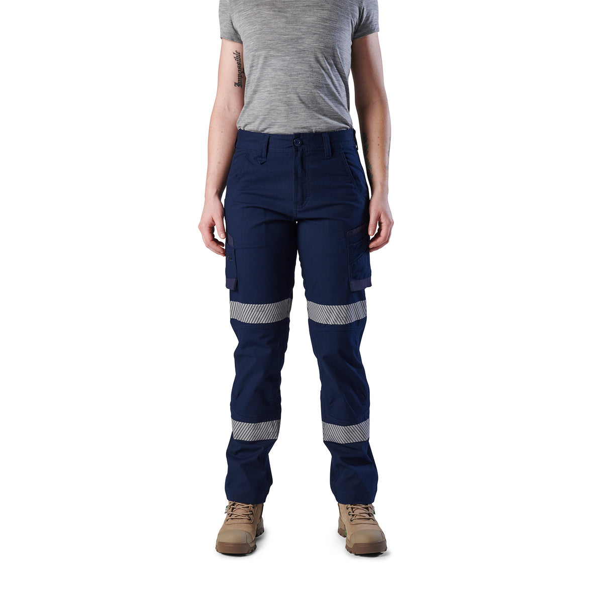 A woman wearing navy blue FXD Women's Taped Cargo Work Pants (WP-7WT) with reflective silver bands on the legs, paired with a grey t-shirt and stone coloured work boots, against a plain white background.