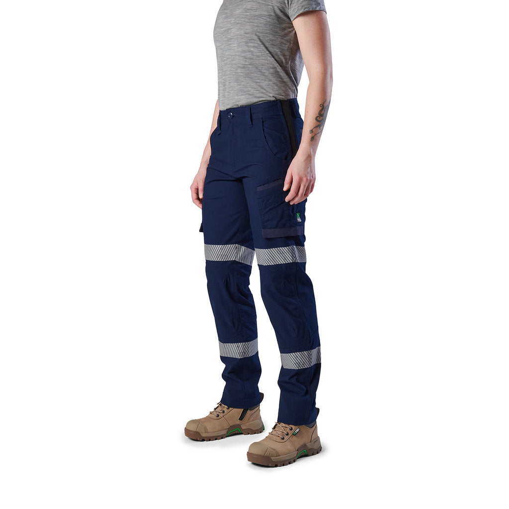 A 3-4 front view of a woman wearing navy blue FXD Women's Taped Cargo Work Pants (WP-7WT) with reflective silver bands on the legs, paired with a grey t-shirt and stone coloured work boots, against a plain white background.
