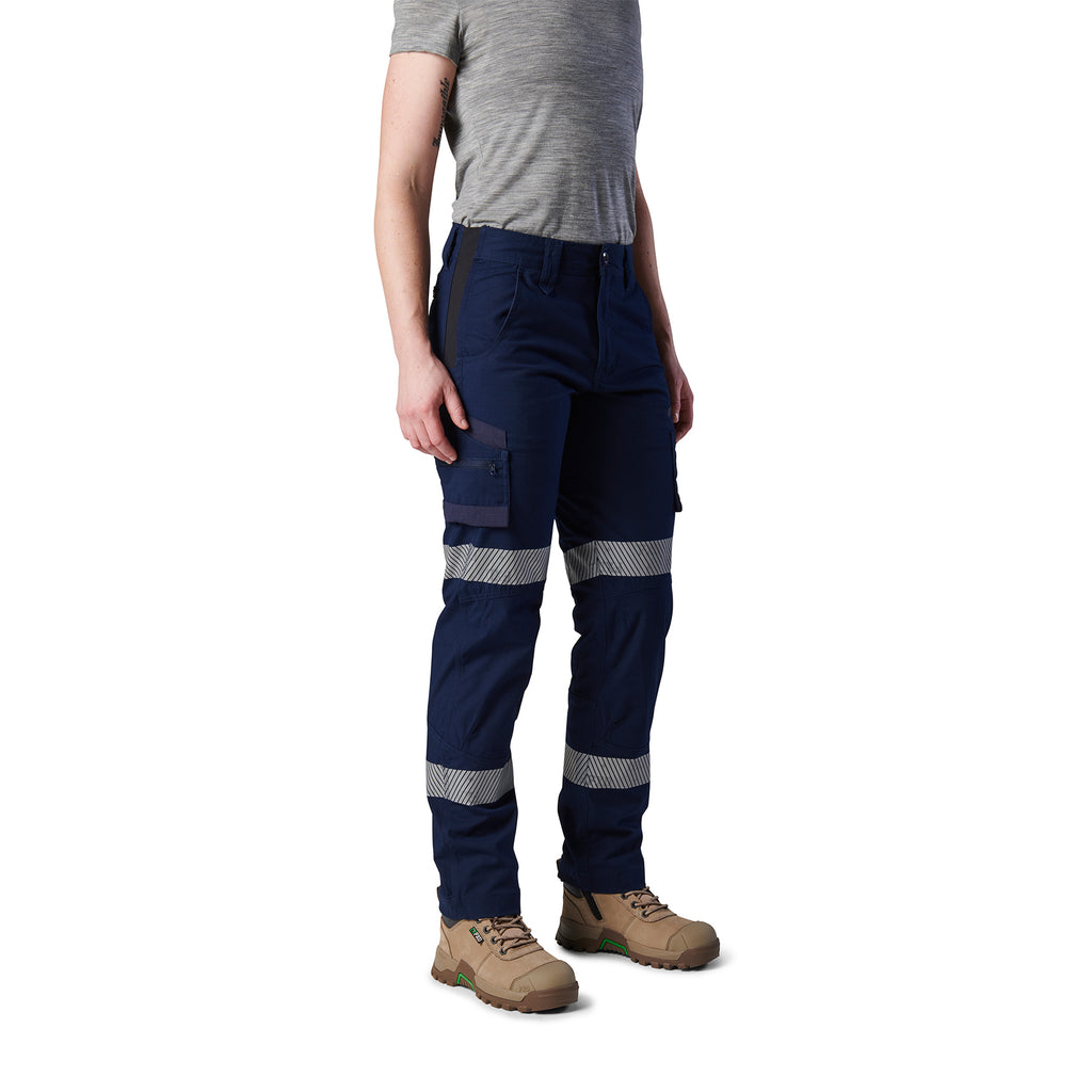 A 3-4 front view of a woman wearing navy blue FXD Women's Taped Cargo Work Pants (WP-7WT) with reflective silver bands on the legs, paired with a grey t-shirt and stone coloured work boots, against a plain white background.