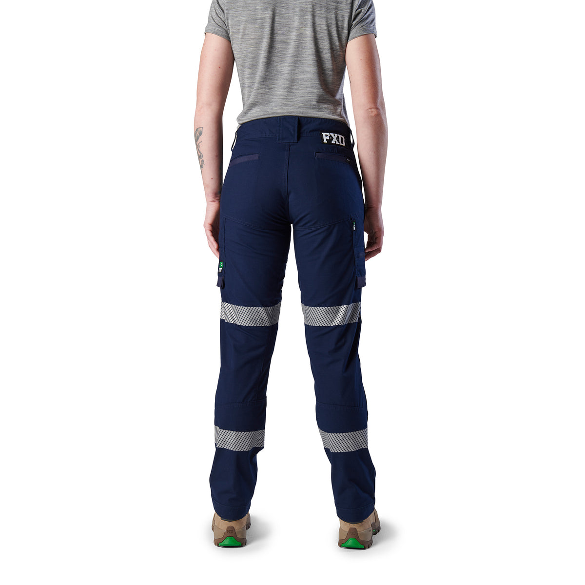 A back view of a woman wearing navy blue FXD Women's Taped Cargo Work Pants (WP-7WT) with reflective silver bands on the legs, paired with a grey t-shirt and stone coloured work boots, against a plain white background.