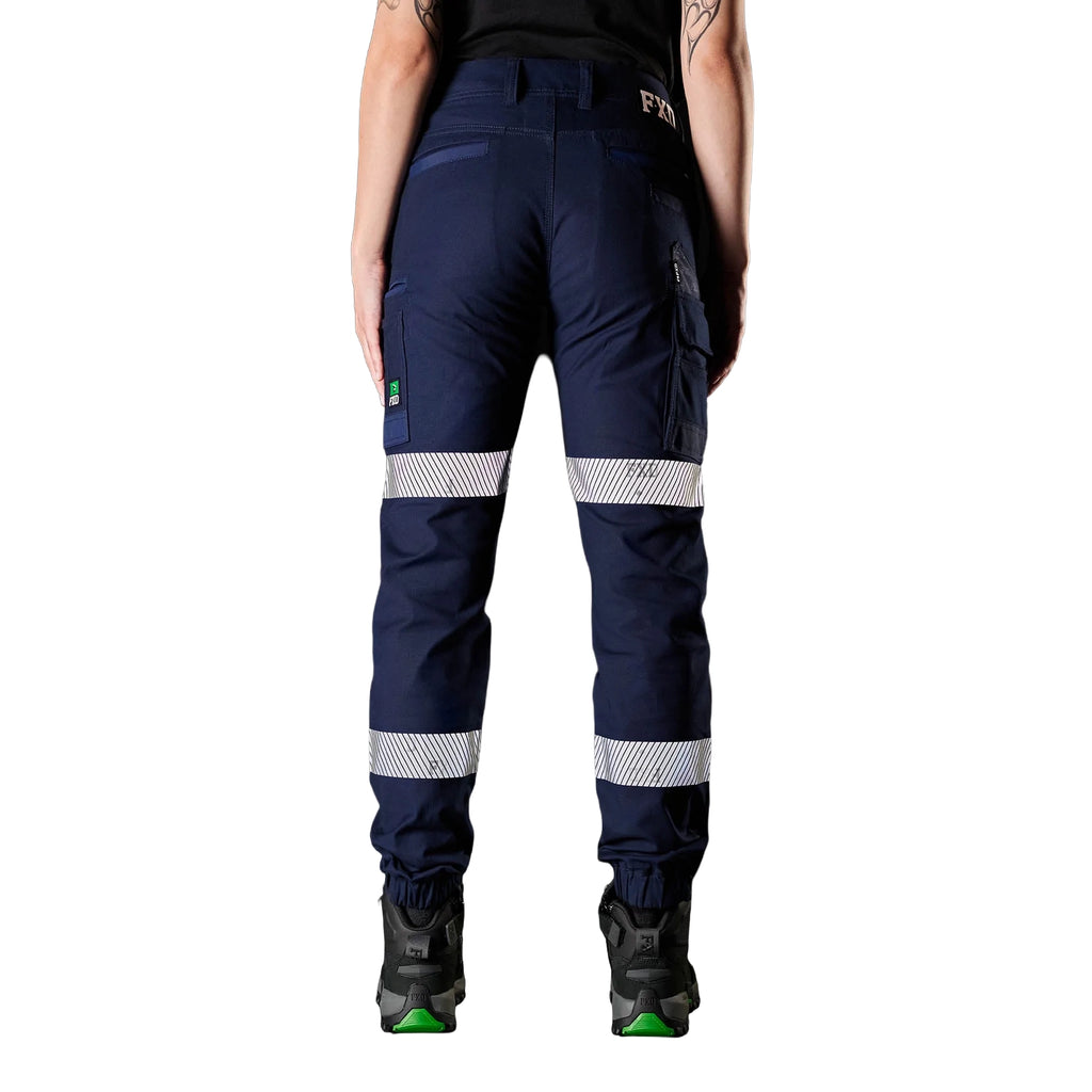 A back view of a woman wearing navy FXD Women's WP-4WT Reflective Cuffed Work Pants (FX12206208) with reflective silver stripes on the thighs and lower legs, paired with black boots against a plain white background.