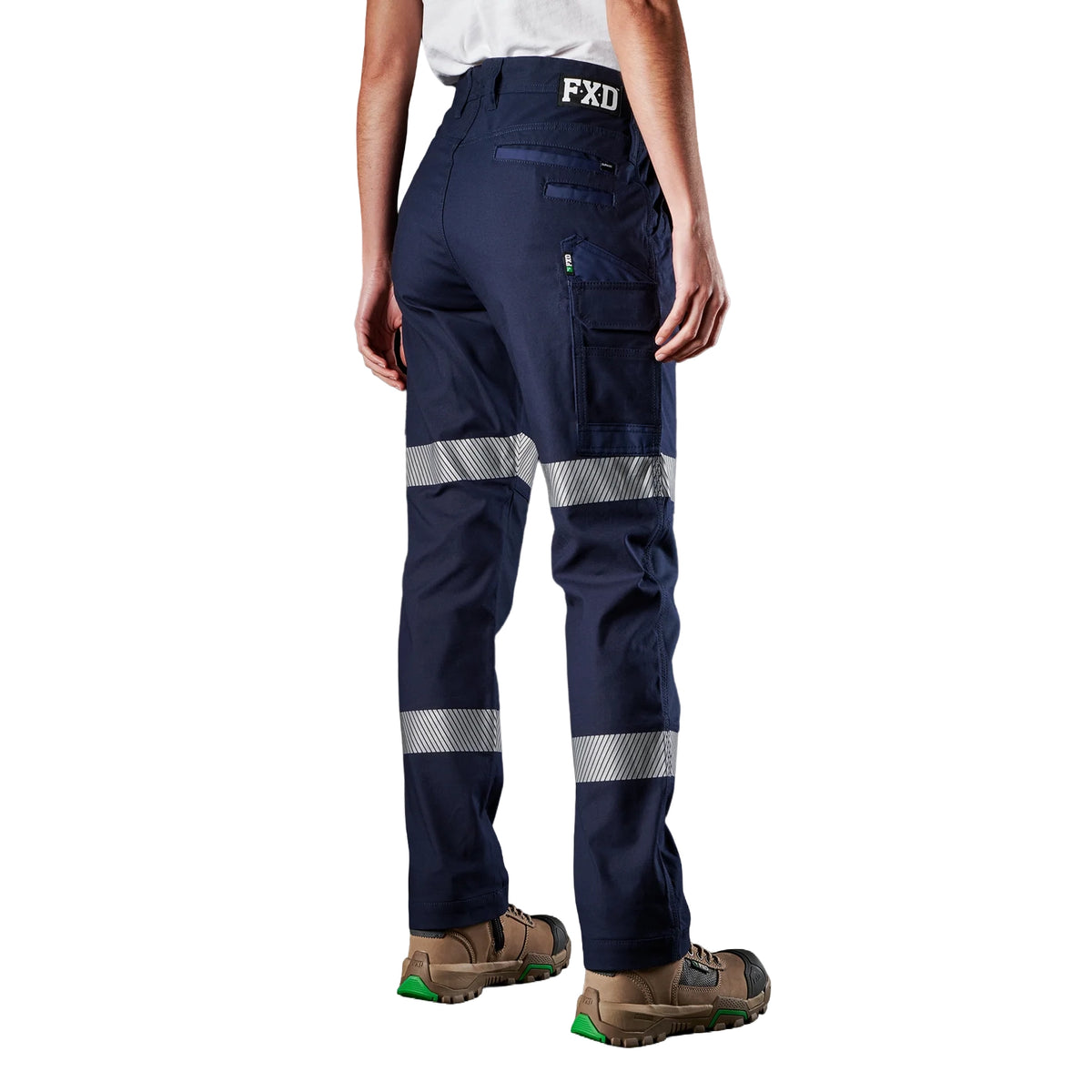 A 3-4 back view of a woman wearing navy FXD Women's WP-4WT Reflective Cuffed Work Pants (FX12206208) with reflective silver stripes on the thighs and lower legs, paired with black boots against a plain white background.