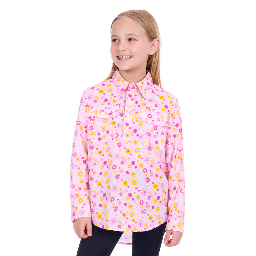 A smiling young girl with long blonde hair wears the Hard Slog Childrens Daisy Half Button L/S Shirt (H6W7101167) in pink, decorated with yellow and pink flowers, paired with dark pants and standing slightly right against a white background.