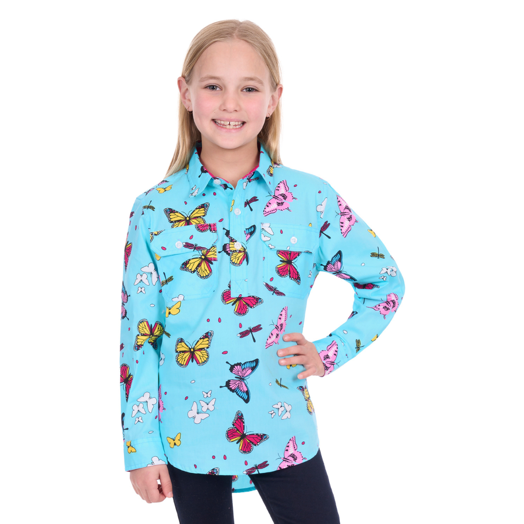 A smiling girl with blonde hair wears the Hard Slog Childrens Iva Half Button L/S Shirt (H6W7101165) in blue by Hard Slog, paired with dark pants, standing against a white background with one hand on her hip.