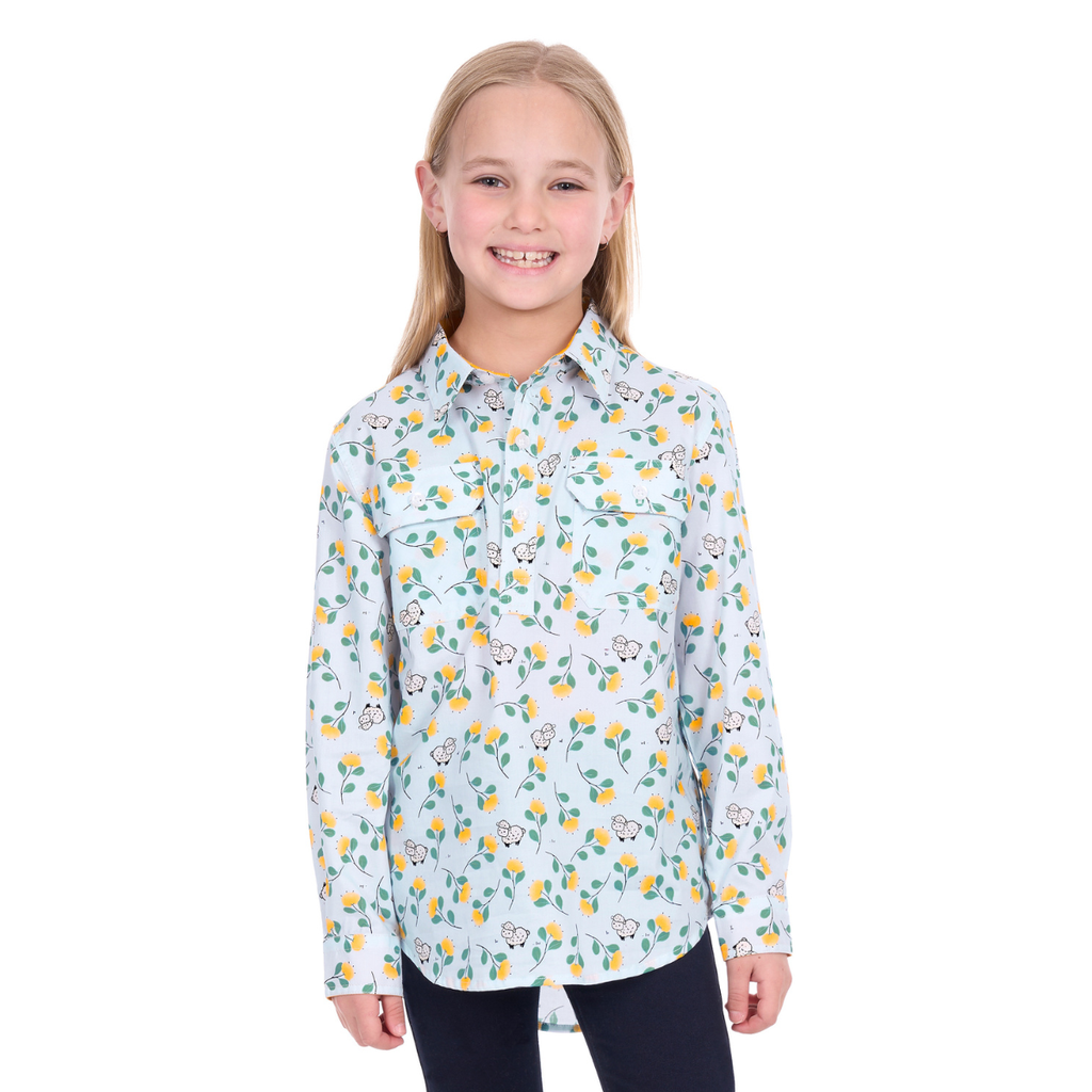 Smiling young girl with straight blond hair wearing the Hard Slog Childrens Suzie Half Button L/S Shirt (H6W7101166) in light blue, paired with black pants, stands against a white background.