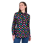 A woman with long brown hair stands smiling, wearing blue jeans and the Hard Slog Womens Rena Half Button Long Sleeve Shirt (H6W2101178) Multi by Hard Slog, featuring colorful polka dots.