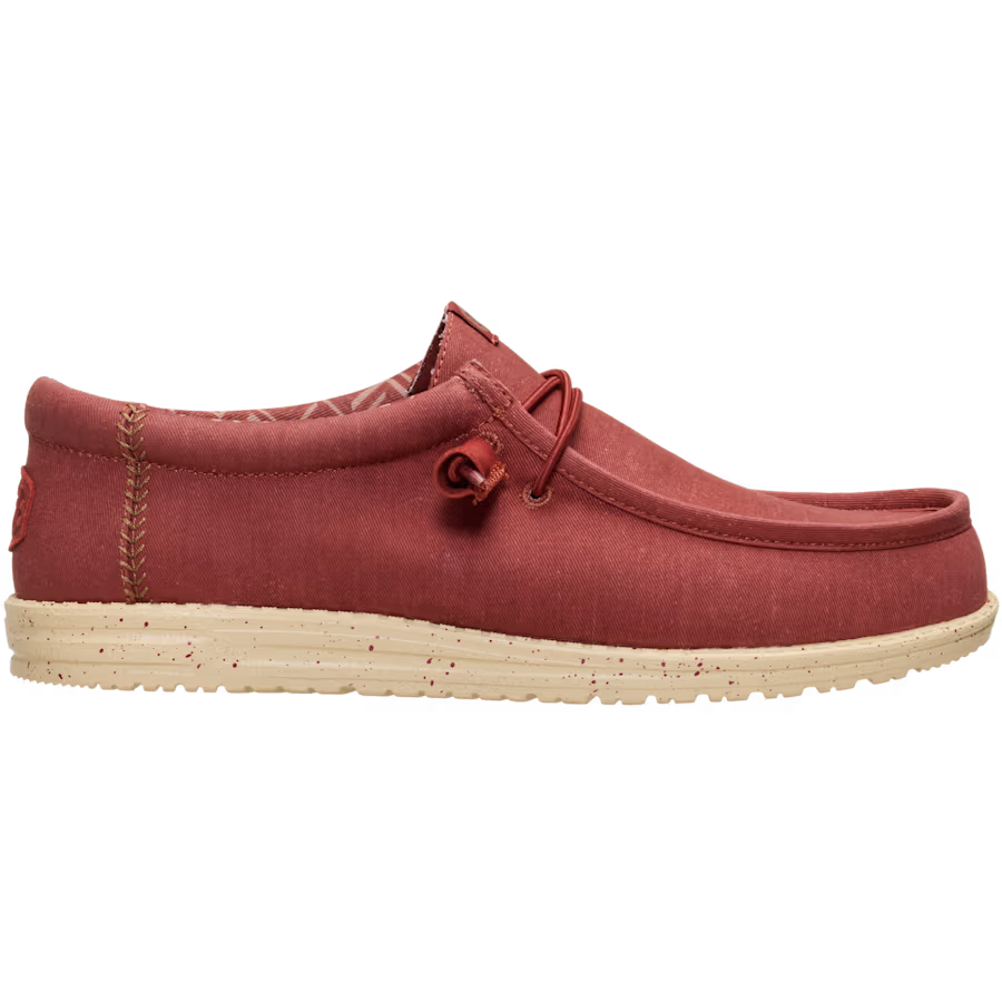 The Hey Dude Mens Wally Stretch Canvas Slip On Shoes in Burnt Red/Boulder Tan feature a red stretch canvas upper, beige speckled sole, visible stitching, cushioned ankle collar, and lace detail near the collar. Brand: Hey Dude.