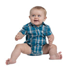 A smiling blonde baby sits on the floor wearing the Just Country Infants Frankie Full Button S/S Jumpsuit (IWSS2627) in Petrol Plaid, featuring short sleeves and a blue and white check pattern, against a white background.