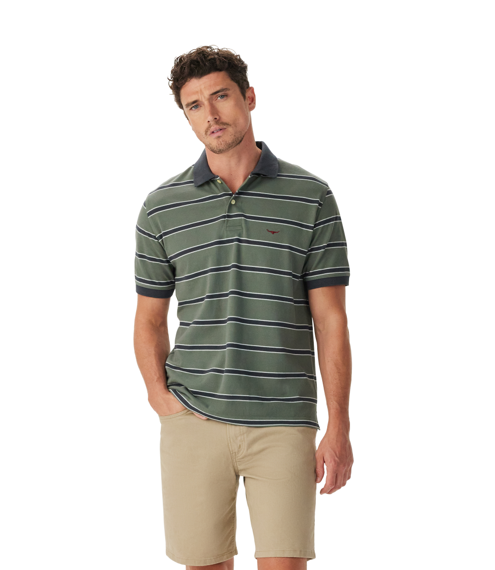 A man wears the R.M.Williams Mens Brunswick Stripe Polo (KP210QS09) in Spruce Green, styled with tan shorts and a dark collar, standing against a white background. Shop online from an official R.M.Williams retailer.