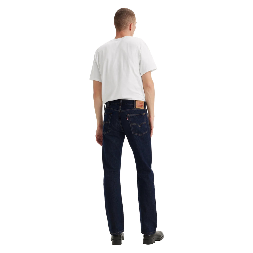 A man with his back to the camera wearing dark blue rinse Levi's Men's 517 Heritage Bootcut Jeans (00517-0216), a white t-shirt, and black shoes, standing against a solid white background.
