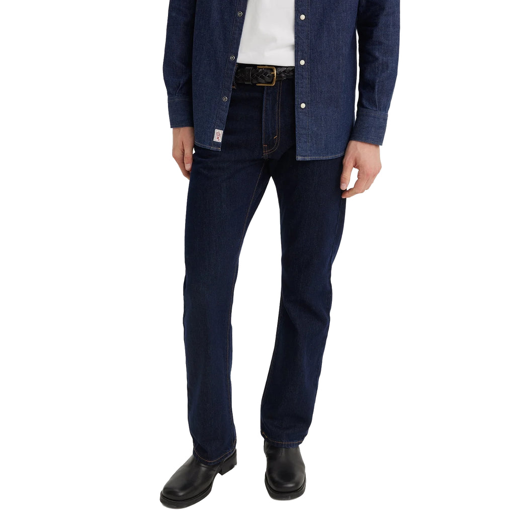 A man from the waist down wearing dark blue rinse Levi's Men's 517 Heritage Bootcut Jeans (00517-0216), a white t-shirt, denim jacket and black shoes, standing against a solid white background.