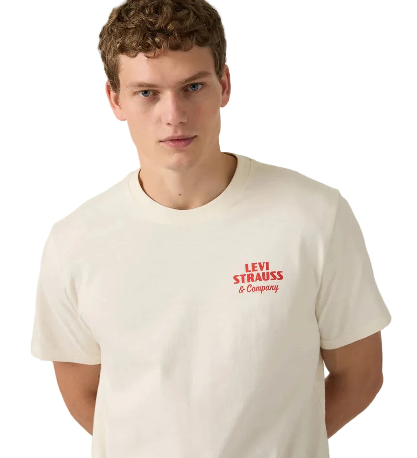 A young man with short curly hair models the Levi's Men's Graphic S/S Tee (16143-2372) Archival Overalls 2 Egret, an off-white relaxed fit tee, against a white background, standing with his arms behind his back.