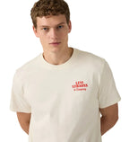 A young man with short curly hair models the Levi's Men's Graphic S/S Tee (16143-2372) Archival Overalls 2 Egret, an off-white relaxed fit tee, against a white background, standing with his arms behind his back.