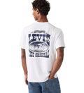 A man with short curly hair wears a Levi's Men's Graphic S/S Tee (16143-2384) Archival Cowboy 1 White—a relaxed fit, white tee with a large blue "LEVI'S THE ORIGINAL" graphic and oval image on the back—paired with blue jeans.