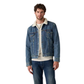 A man wears a Levi's Men's Type 3 Sherpa Trucker Denim Jacket (16365-0292) In The Morning over a light shirt and blue jeans, standing against a plain white background.
