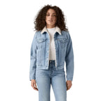 A woman with curly hair wears the Levi's Womens Original Sherpa Trucker Denim Jacket (36136-0071) Feeling Everything—light blue denim with a white sherpa collar—over a white top and blue jeans, standing against a plain white background.