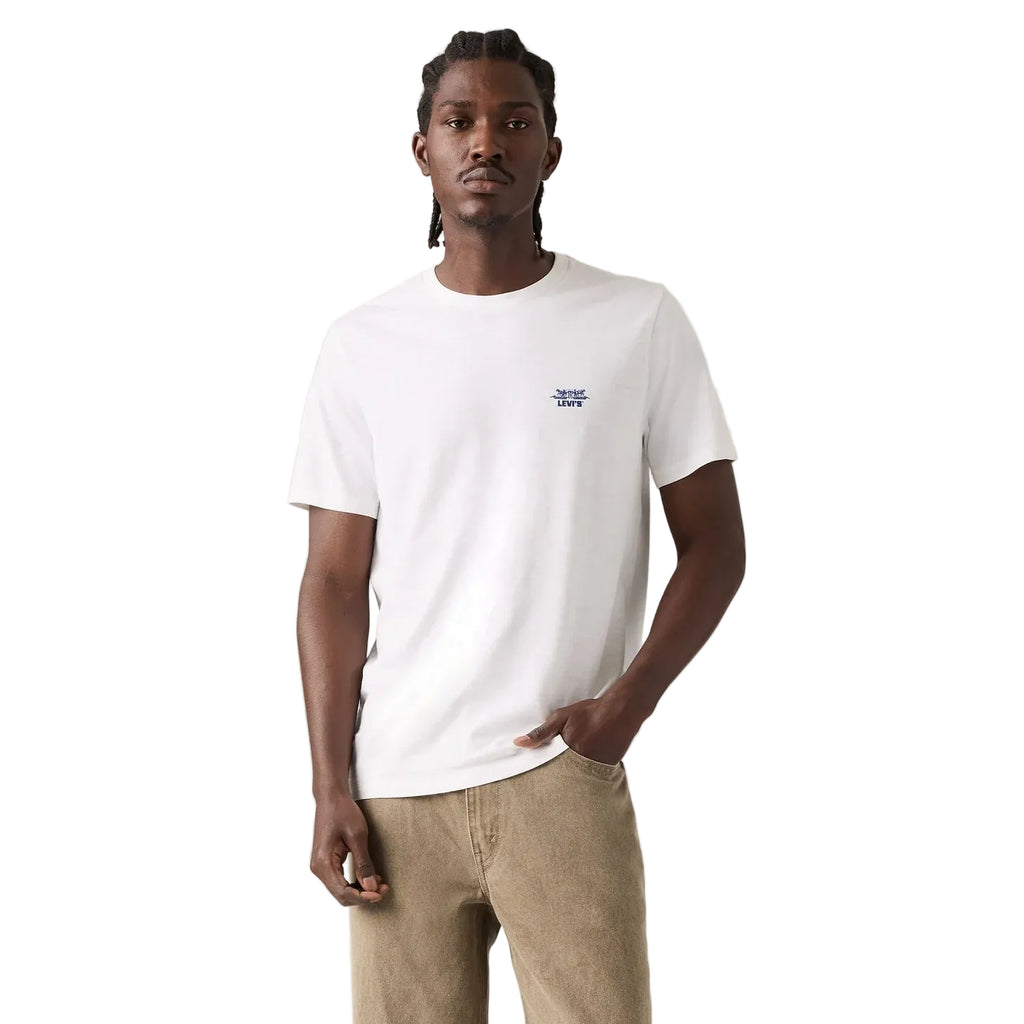 A man wearing a white crew-neck t-shirt with a small blue embroidered logo on the left chest, paired with beige trousers, standing against a plain light white studio background.