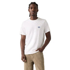 A man wearing a white crew-neck t-shirt with a small blue embroidered logo on the left chest, paired with beige trousers, standing against a plain light white studio background.