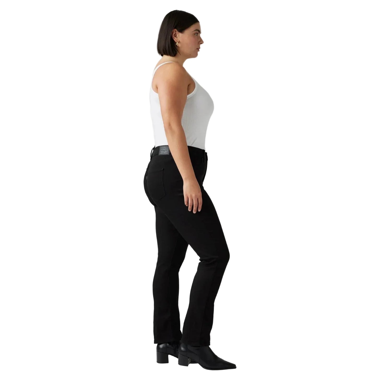 A woman in side profile wearing a white tank top, black pants, and black ankle boots, standing against a white studio background.
