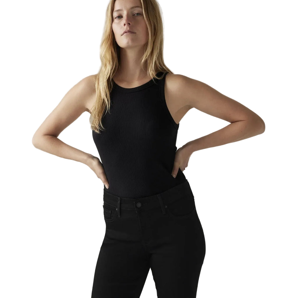 A blonde model wearing a black ribbed sleeveless tank top and black jeans, posing with hands on hips against a white studio background.