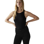 A blonde model wearing a black ribbed sleeveless tank top and black jeans, posing with hands on hips against a white studio background.