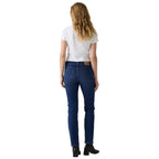 A woman viewed from behind wearing dark blue jeans, a white t-shirt, and black ankle boots against a white studio background.