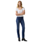 A woman modeling a pair of dark blue  jeans with a white short-sleeve top and black ankle boots, standing against a white studio background.